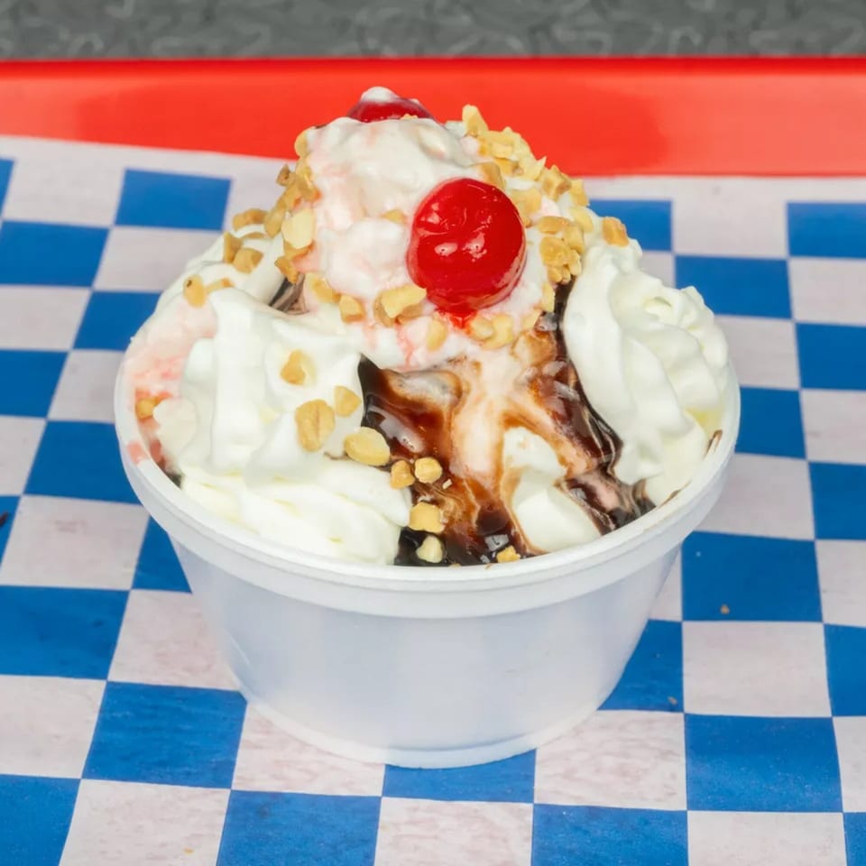 Ice cream sundae with whipped cream, chocolate syrup, nuts, and cherries in a white cup.