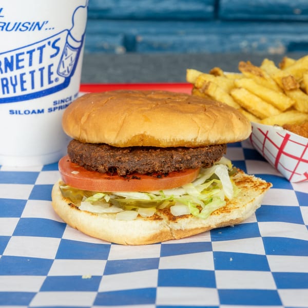 A hamburger with lettuce, tomato, and pickles, alongside fries and a drink on a checkered paper.