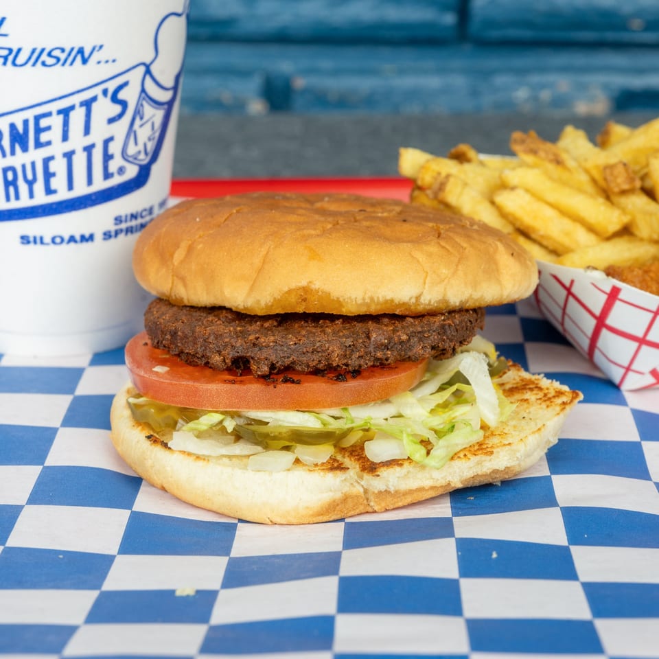 A hamburger with lettuce, tomato, and pickles, alongside fries and a drink on a checkered paper.