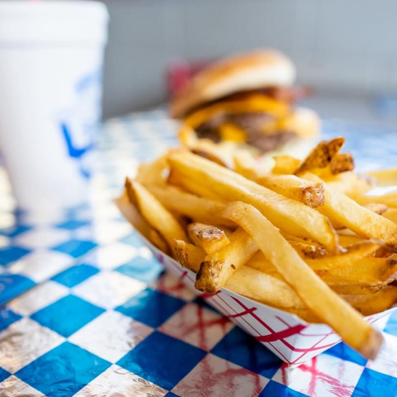Golden french fries in a paper tray, with a blurred burger and drink on a blue checkered table.
