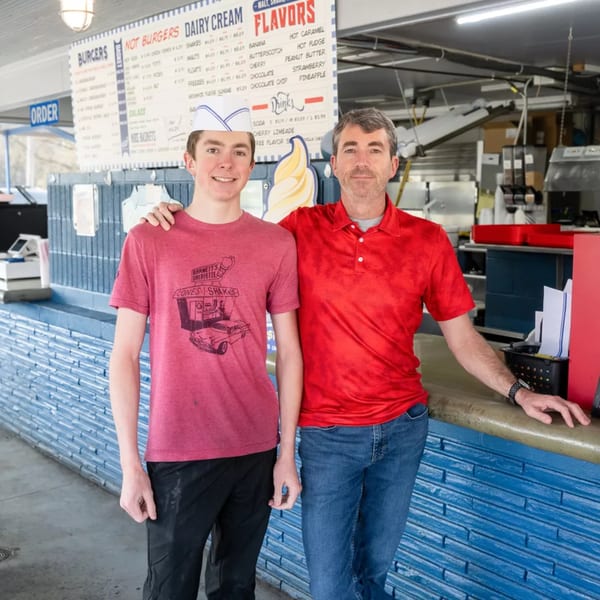 Two men, one in a paper hat, stand in front of Barnett's Dairyette menu board.