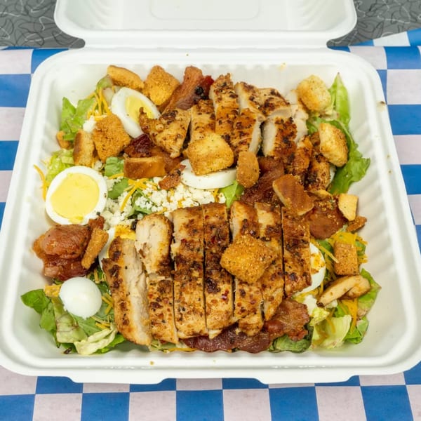 Grilled chicken salad with hard-boiled eggs, croutons, and cheese in a white takeout container.
